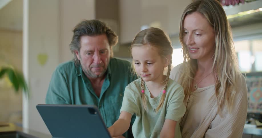 Father with beard and turquoise shirt and blonde mother wearing brown dress help daughter with braids use tablet in kitchen at home
