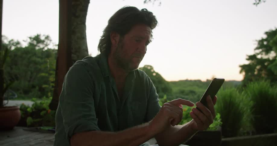 Man smiles while sitting outdoors scrolling on his phone