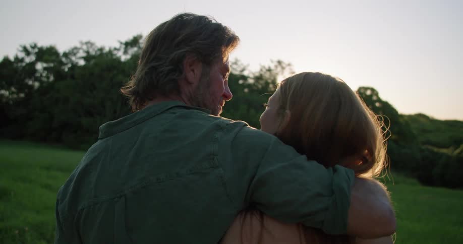 A man and woman stand with their arms around one another watching the sunset and he leans over and kisses her