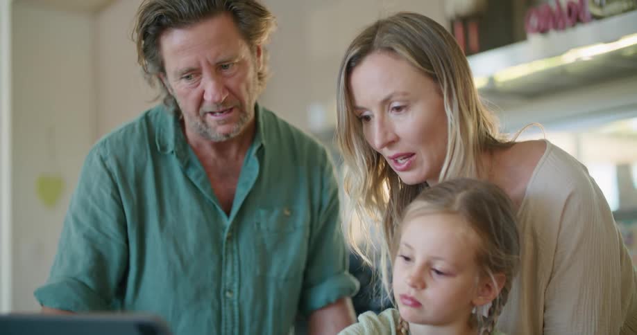 Father with beard and turquoise shirt and blonde mother wearing brown dress help daughter with braids use tablet in kitchen at home