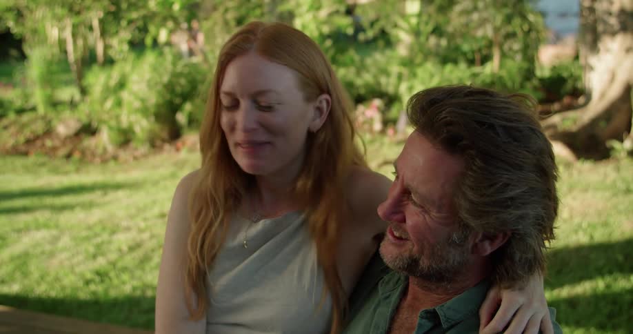 Man with beard and blue shirt holds woman with red hair and grey vest sitting in garden lovingly looking into each others eyes