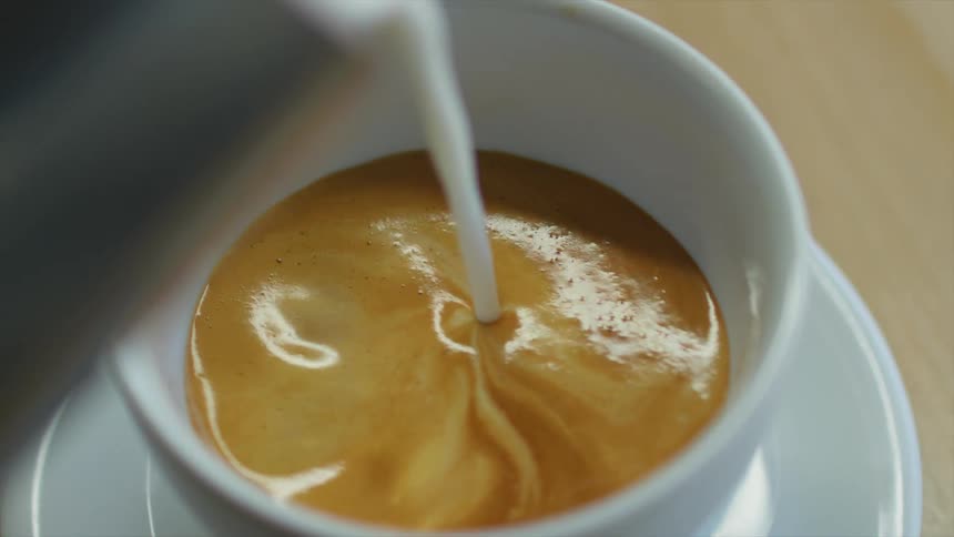 Milk pouring on coffee inside a white porcelain cup over a table