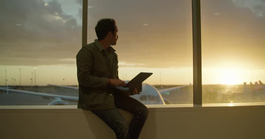 A man uses a laptop while sitting on a window ledge at an airport at sunset