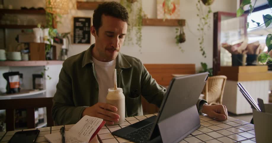 Man with moustache green jacket and white shirt looks at tablet and takes notes in notebook drinking beverage in cafe