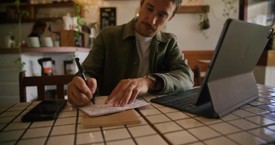A man taps his pen while lost in thought for a moment before writing in a notebook while sitting at a table in a cafe during the day