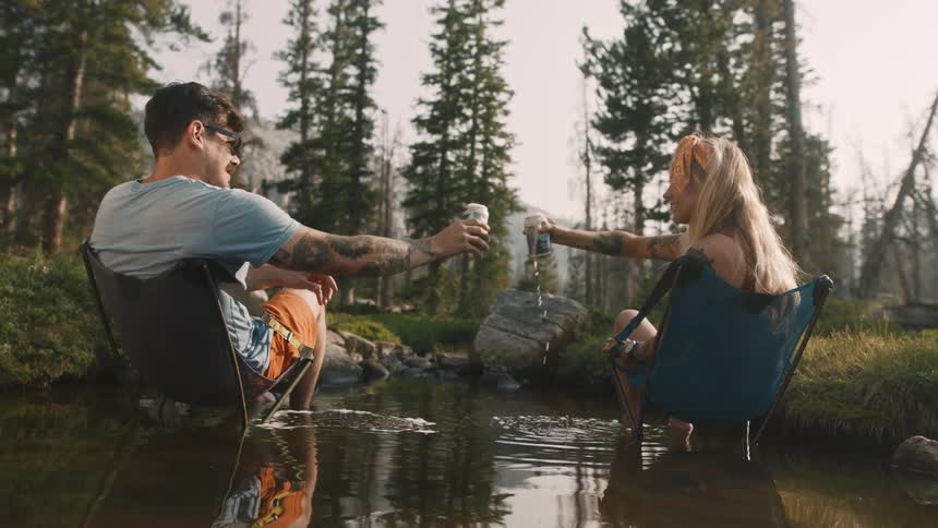 A man and woman open beers and toast while sitting in the stream