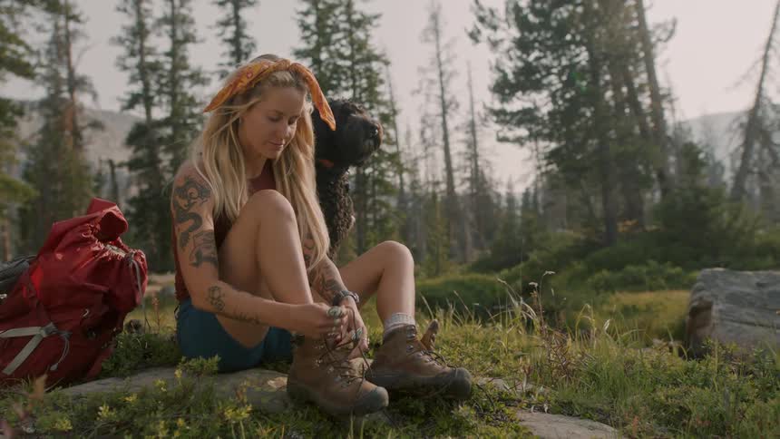 Dog sits near woman unlacing her hiking boots with mountains in the background