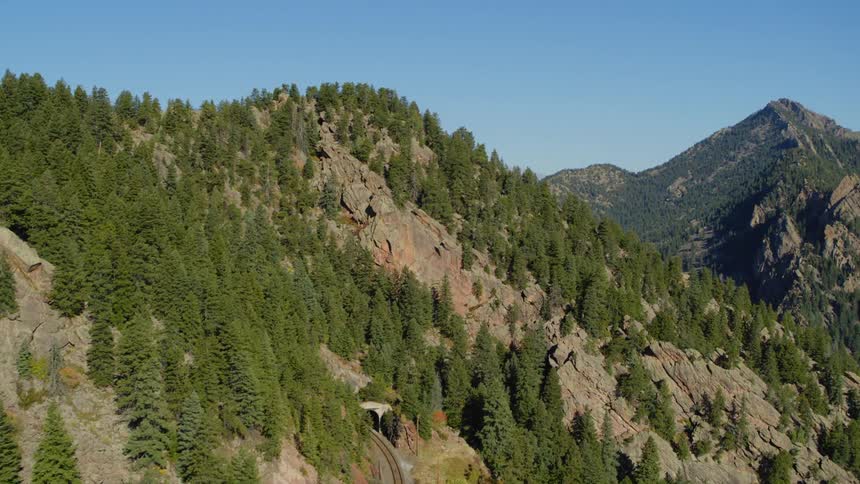 Train tracks disappear into a mountain tunnel with tree covered peaks under a blue sky