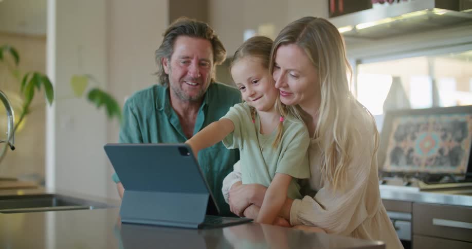 Father with beard and blue shirtandblonde mother wearing brown dress helps daughter with braids use tablet in kitchen at home