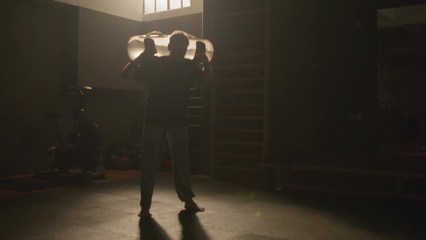Man performs squats with water weight container on his back in dim gym