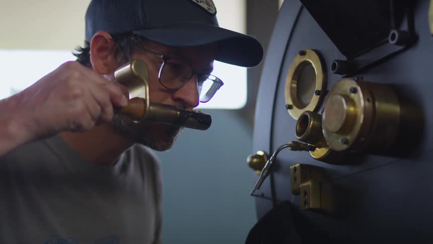 Man wearing glasses and baseball cap sampling roasted coffee beans from a working oven