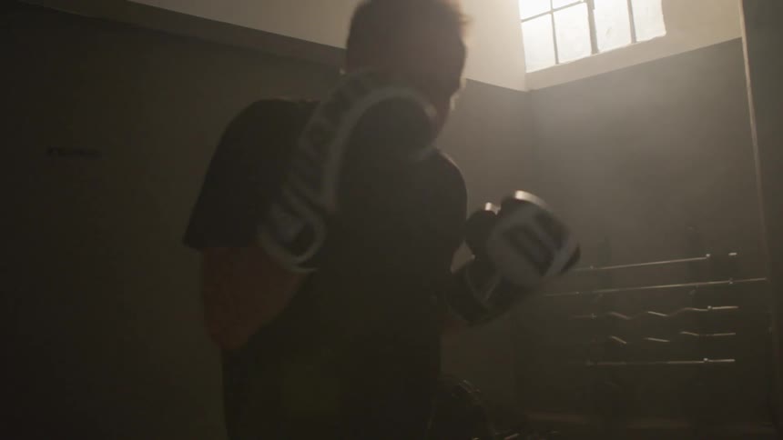Sunlight filters through a window over a man shadow boxing on a dimly lit gym at morning