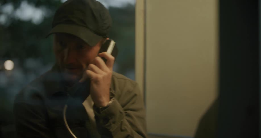 Man wearing baseball cap and jacket places a call on a public phone