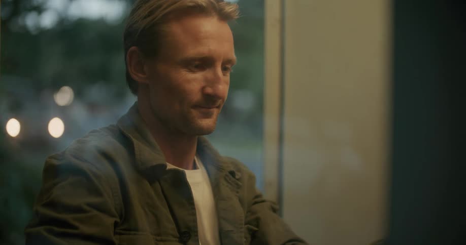 A man is standing inside in front of a window talking on a telephone and he hangs up and stands thinking
