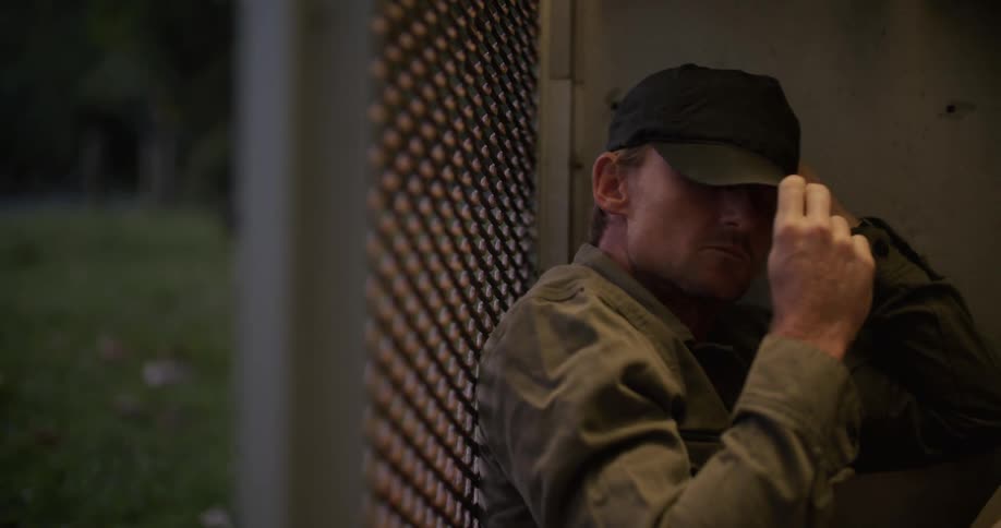 Man puts on hat and draws coat close as he tries to sleep in mesh sided enclosure as animals move around in the background near the woods