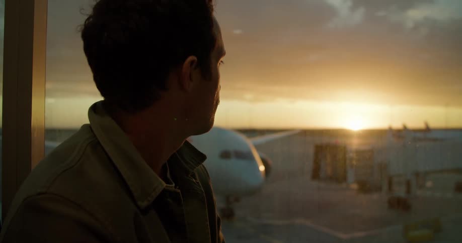 A man types on his laptop inside an airport as he looks out the window at a large plane