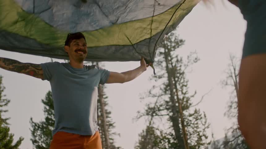 A man laughs as him and a woman outdoors lift a parachute over their heads and let it fall on them