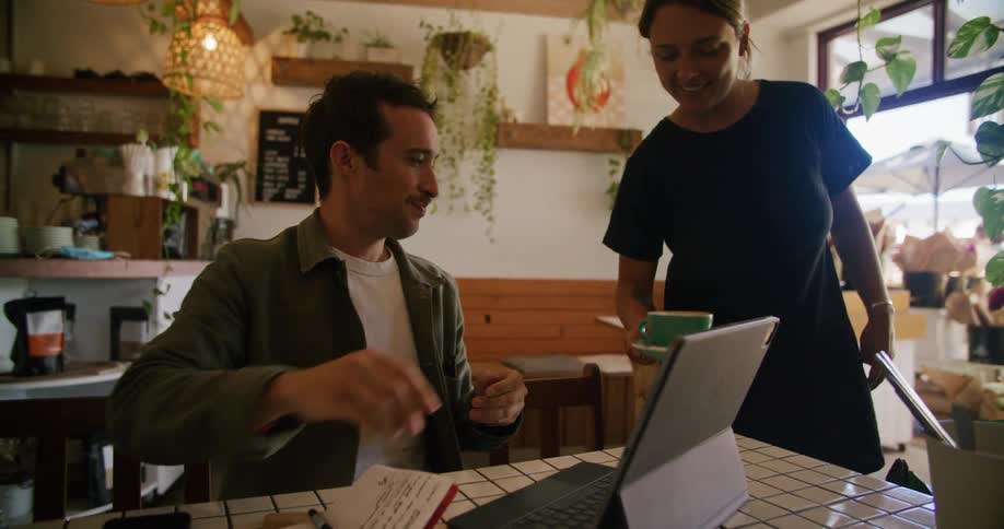 Man sitting on a table at a restaurant working on a laptop is served a cup of coffee
