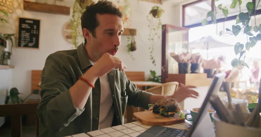 A man takes a bite from his sandwich while gazing at his laptop at a table in a cafe during the day