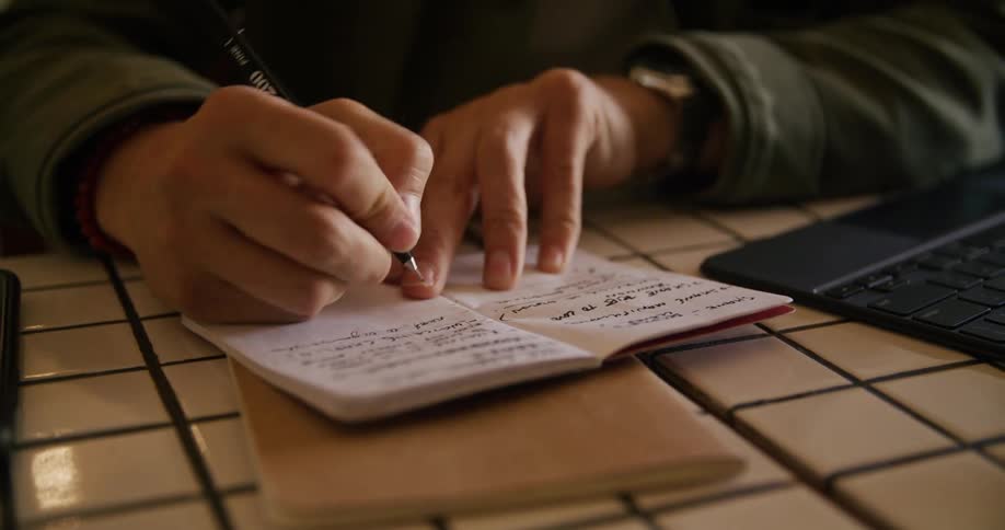 A man is writing in his notebook by the laptop
