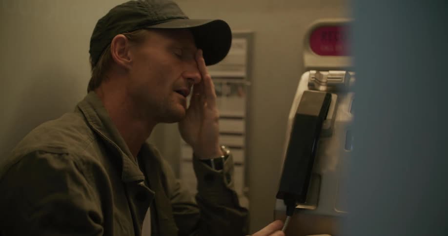 A man hangs up a payphone takes off his cap and rubs his face in frustration