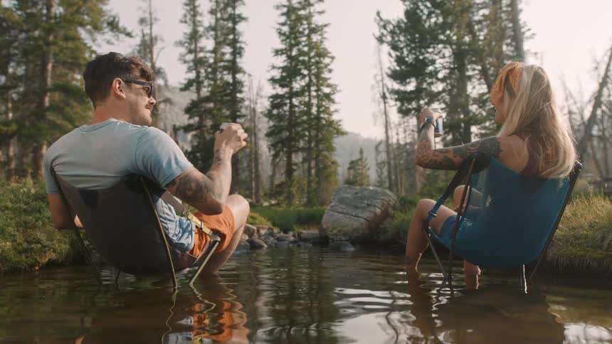 Man and woman sitting in chairs set in edge of pondnear forest toast each other