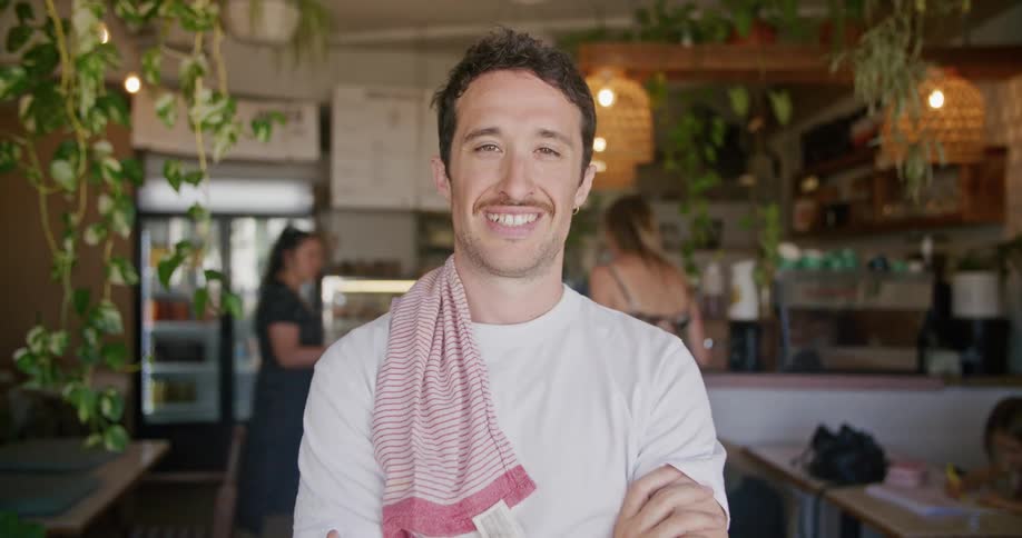 Man with moustache white t shirt faces the camera with eye contact smiling in cafe