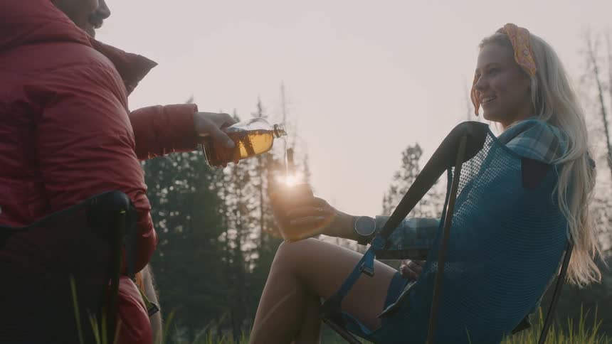 Man pours woman a drink as they sit in chairs facing conifer forest