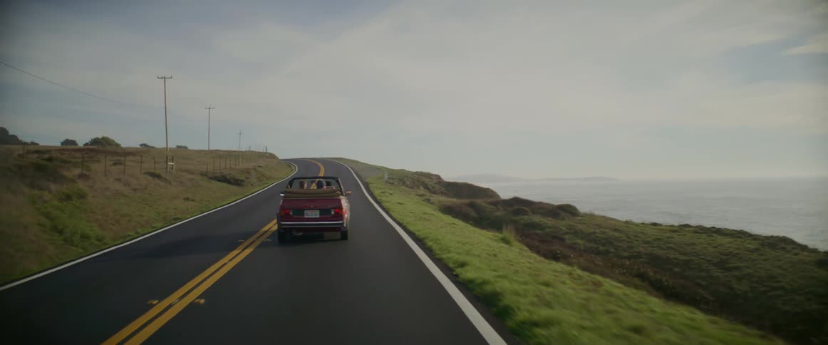 People drive along highway in convertible between fenced field and cliff