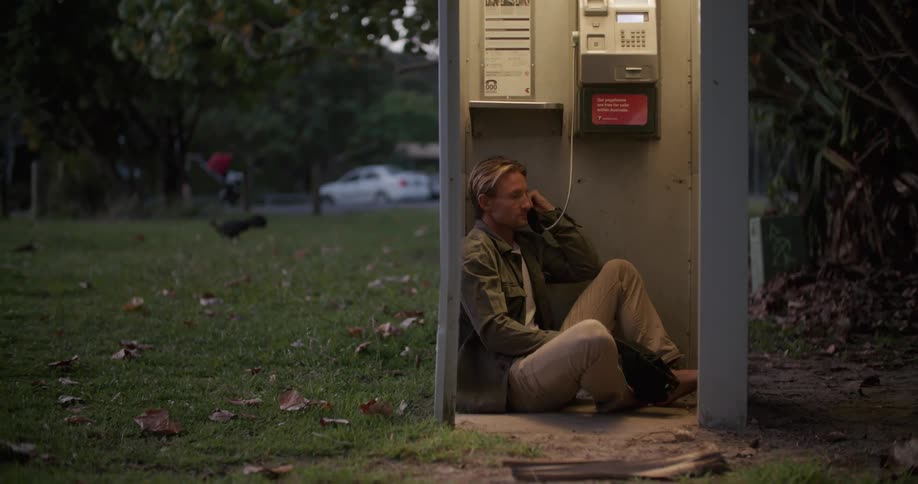 A man sits on the floor of a phone booth and talks on the phone as people walk past in the distance