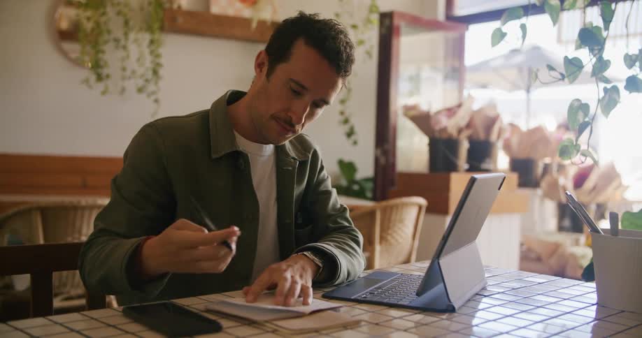 Man sits at table in store making notes from computer as pedestrians pass