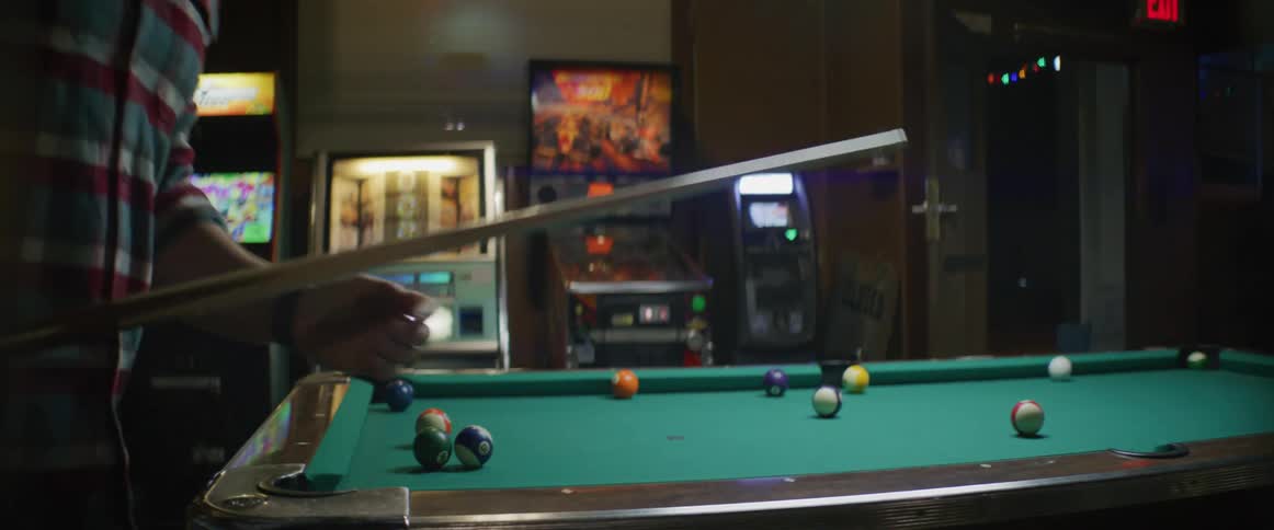 Man wearing knit hat plays pool in room near arcade games and slot machines
