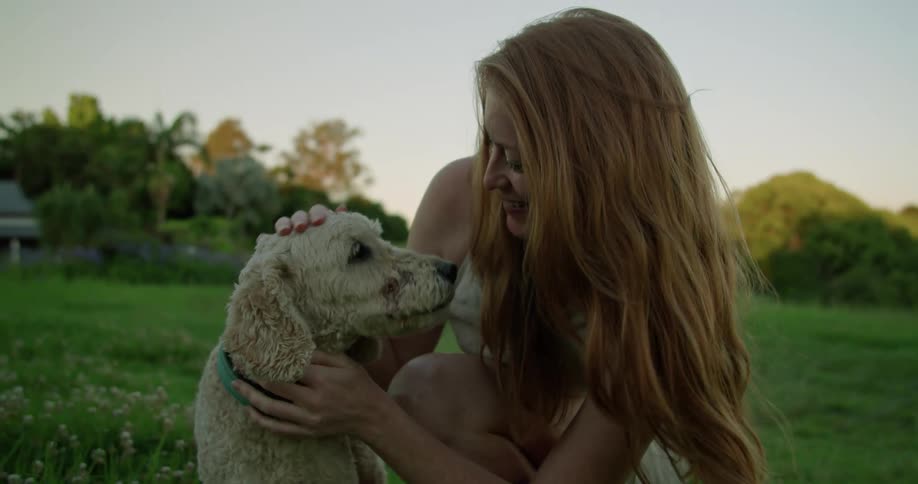 Red haired woman cuddles and pets wire haired dog as they crouch in field with house in background