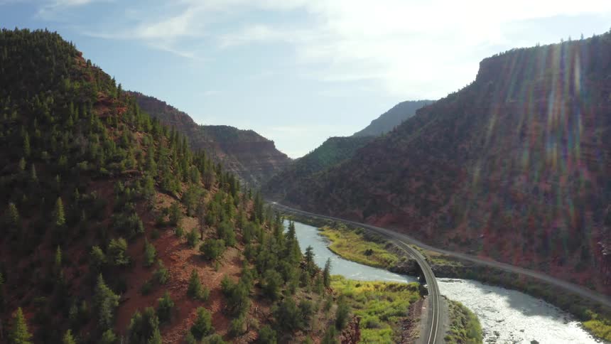 Bright sun shines into a valley where a light blue river flows under a modern road situated between forested mountains