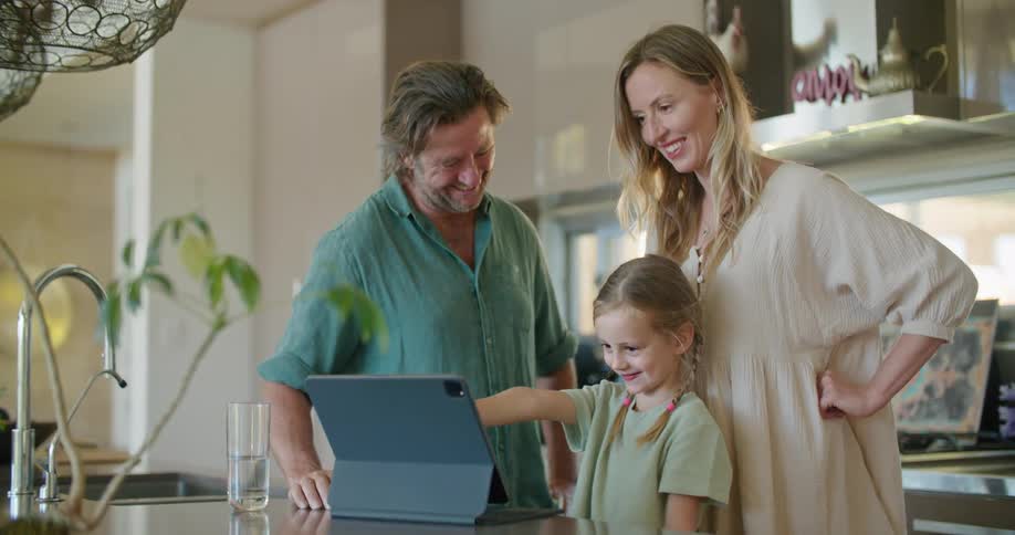 Father with beard blue shirt daughter with braids and blonde mother wearing brown dress use and look at tablet in kitchen at home