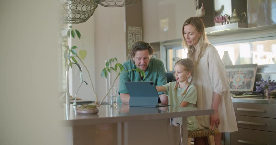 A man and woman stand behind a girl as she uses a computer tablet in a stand on a kitchen counter
