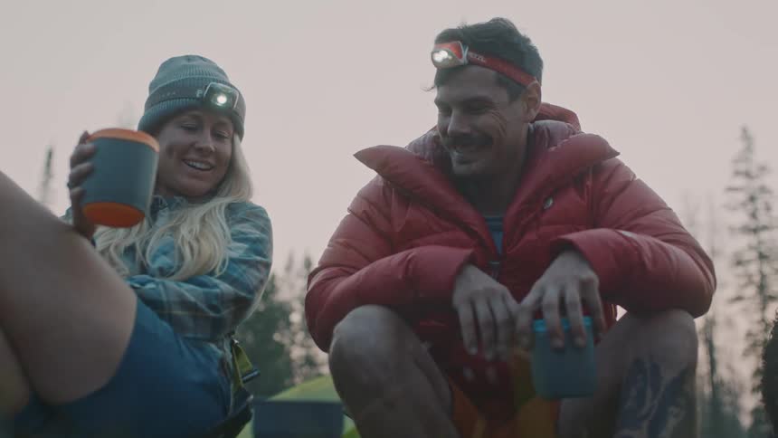 A man and woman wearing headlamps sitting near a fire at their campsite talking and smiling
