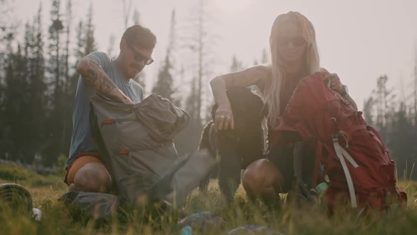 A dog stands between a man and woman as they unpack their backpacks to set up their camp site