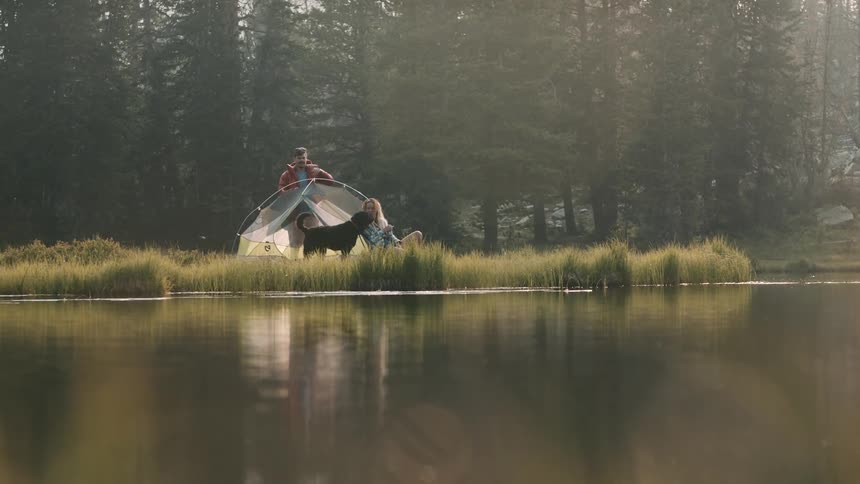 A man stands behind a tent on the shore as a dog goes up and licks the face of a woman sitting in front of the tent