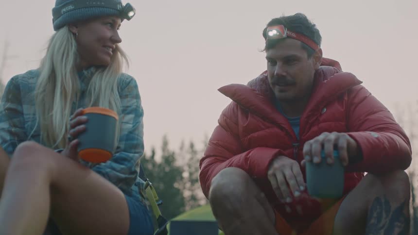 Young couple wearing headlamps sitting in the forest drinking hot beverages at evening