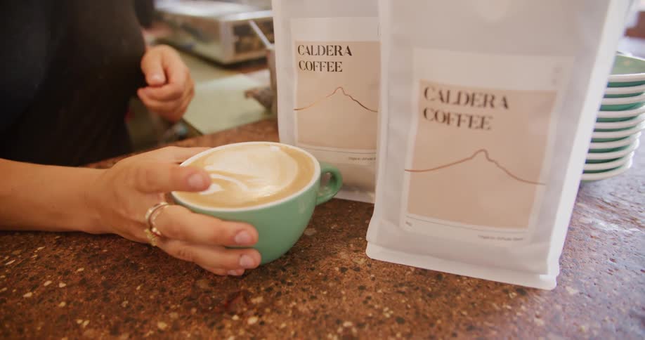 Baristas hand setting down a latte in front of coffee bags
