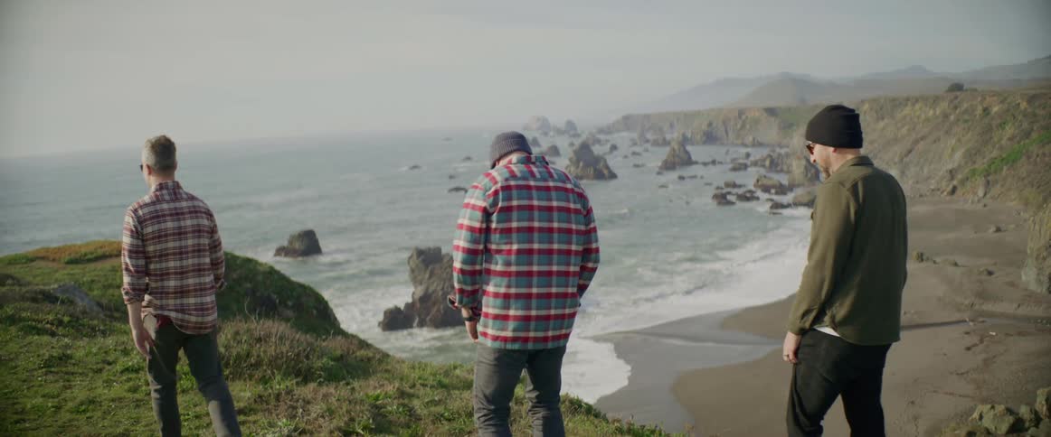 Three men with beanies plaid shirts and sunglasses wanders on cliff overlooking beach with big rocks
