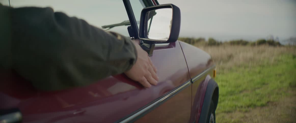A person sitting in the passenger seat of a car parked in grassland taps their hand rhythmically on the outside of the passenger side door