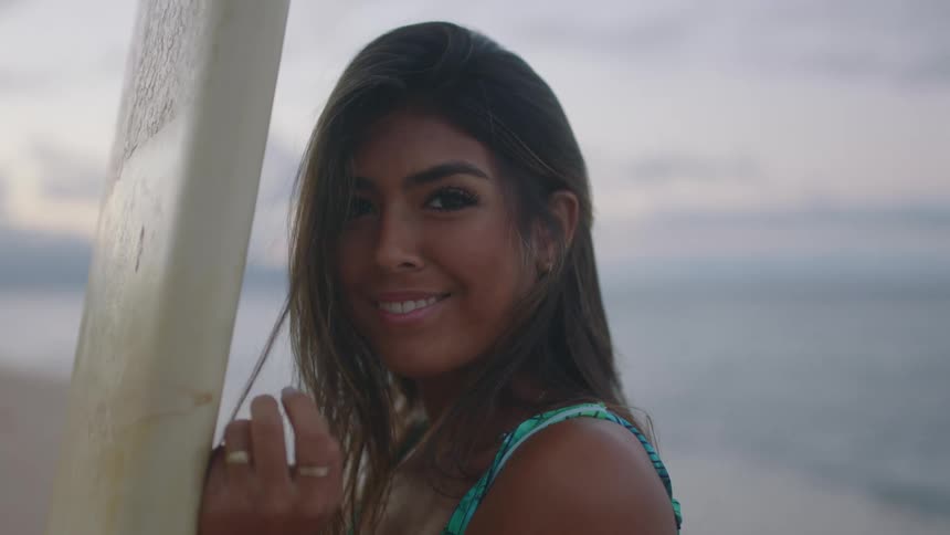 Woman with wavy dark brown hair and leaf print turquoise swimming costume holds surfboard looking at camera smiling on the beach at dusk