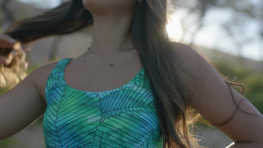 Woman with wavy dark brown hair and leaf print turquoise swimming costume touches her hair looking around as the sun shines in the background on the beach