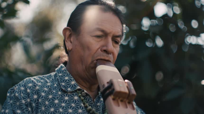 A native american man plays a traditional wind instrument while standing outdoors during the day