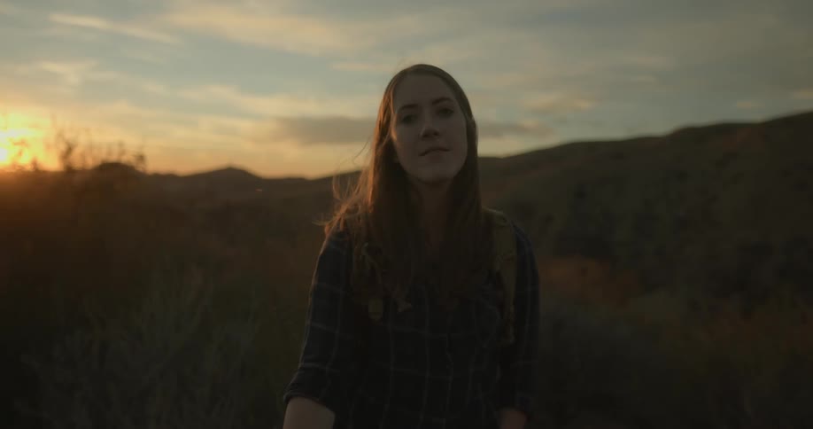 Woman stands in field near hills back lit by sun sitting on the horizon in a red sky
