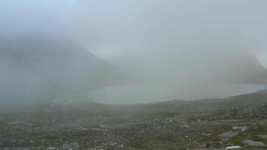 Aerial view of a white car parked on a clearing near a lake under the mist close to the mountains