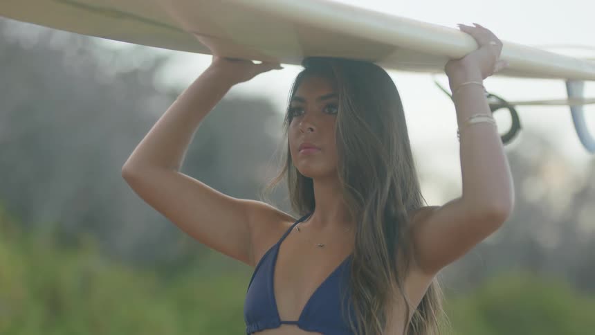 Woman with brown wavy hair and dark blue bikini holds surfboard on top of her head