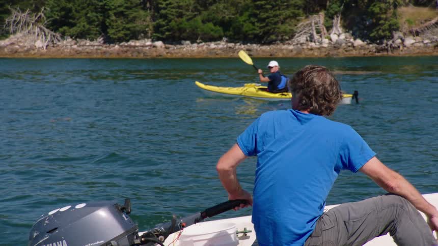 Man pilots boat and and waves at two people kayaking with homes visible in background